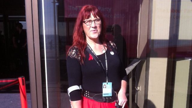A white woman with red hair and glasses, wearing a black top and a red skirt and using a crutch, stands outside a conference centre